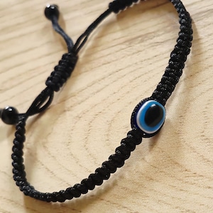 Può includere: Un braccialetto in corda intrecciata nera con una perla centrale a forma di occhio di Allah con cerchi concentrici blu, bianchi e neri. Il braccialetto ha una chiusura a nodo scorrevole regolabile con due piccole perle nere e il testo Symoru.