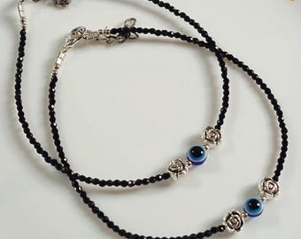 May include: Two black beaded anklets with silver-toned floral accents and blue evil eye charms. The anklets are displayed on a white surface with floral details. The text "Symoru" is in the top left corner.