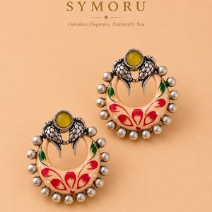 May include: A pair of circular earrings featuring a cream-colored base with red and green floral patterns. Each earring is accented with a yellow stone at the top, two silver bird figures, and a border of small silver beads. The text SYMORU is visible.