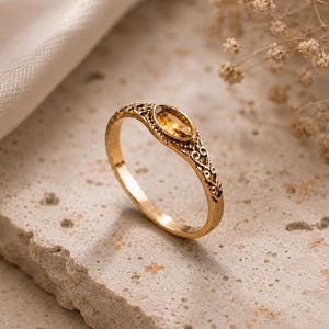 May include: A gold-toned metal ring featuring a marquise-cut amber-colored gemstone set in the center. The band displays an intricate, raised filigree pattern along the sides, extending from the central stone setting to the rest of the ring.