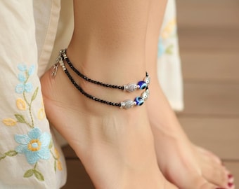 May include: Black beaded anklets with silver-toned fish charms and blue evil eye beads. The anklets are worn on the ankle. The toenails are painted red. The background is a wooden surface.