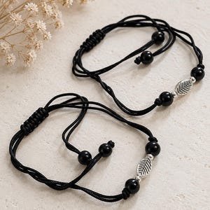 Op de afbeelding: Twee verstelbare zwarte koordarmbanden met een centrale zilverkleurige metalen bladbedel geflankeerd door twee gladde zwarte kralen. Elke armband is voorzien van zwarte kralen aan de uiteinden en een schuifknoopsluiting.