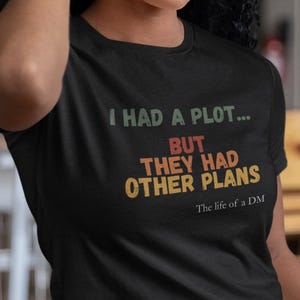 May include: Black t-shirt with the text "I HAD A PLOT... BUT THEY HAD OTHER PLANS The life of a DM" in a retro font. The shirt is worn by a person with curly dark hair and gold hoop earrings.