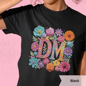 May include: Black t-shirt featuring a floral design with the letters "DM" in a colorful, decorative font. The design includes various dice and flowers in shades of blue, purple, orange, and pink. The shirt is a crew neck.