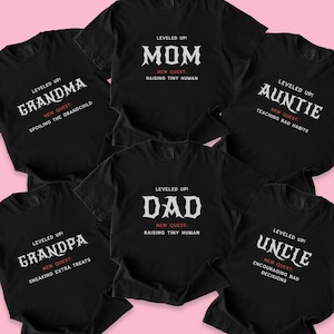 May include: Black t-shirts with white and red text. The shirts say "Grandma", "Mom", "Auntie", "Grandpa", "Dad", and "Uncle". Each shirt has the words "Leveled Up!" and "New Quest" printed on it.