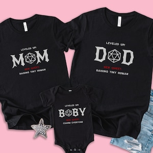 May include: Black t-shirts and a onesie with white and red text. The shirts say "MOM" and "DAD" with a geometric design, and the onesie says "BOBY". All shirts have the text "RAISING TINY HUMAN" or "CHARM EVERYONE".