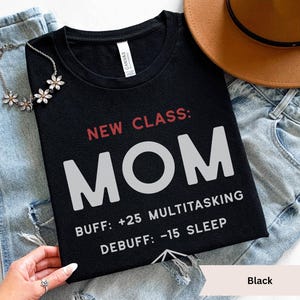 Puede incluir: Una taza de cerámica blanca con asa negra y el texto "But First, Coffee" en negrita negra. La taza se encuentra sobre un fondo borroso, sugiriendo un ambiente acogedor.