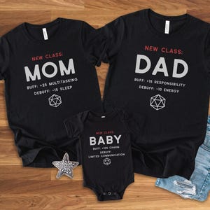 Può includere: T-shirt nere e un body per neonati con testo bianco. Le magliette riportano "MOM" e "DAD", e il body dice "BABY". Ogni articolo ha un elenco di statistiche "BUFF" e "DEBUFF". Sono visibili anche una stella e una forma geometrica.