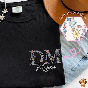 May include: A black t-shirt featuring the letters DM filled with a floral pattern and the name Megan written below in white script. A small hexagonal graphic shows a floral pattern with dice, and a silver flower necklace rests on the collar.