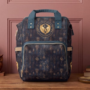 Könnte beinhalten: Ein dunkelblauer Rucksack mit einem sich wiederholenden goldfarbenen Muster aus stilisierten Schwertern und floralen Motiven. Auf der Vorderseite befindet sich ein kreisförmiges Logo mit dem Text Little Leaf's Adventure Bag, zwei Tragegriffe und eine große Reißverschlusstasche.
