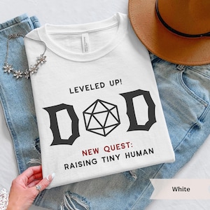 May include: White t-shirt with the text "LEVELED UP! D&D NEW QUEST: RAISING TINY HUMAN" in black. The shirt features a Dungeons & Dragons die graphic. The shirt is displayed with blue jeans and a brown hat.