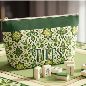 May include: A zippered pouch featuring a green and cream floral pattern with the word TILES embroidered in green. The pouch is accompanied by several white rectangular mahjong tiles with green and red markings, arranged in front of the bag.