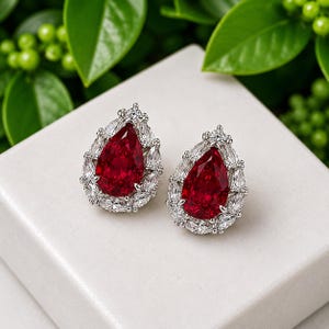 Può includere: Un paio di orecchini a lobo con una gemma rossa centrale a forma di pera, circondata da un alone di piccole pietre chiare taglio marquise incastonate in una montatura in metallo color argento su una superficie bianca.