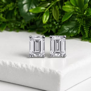 Puede incluir: Un par de pendientes de botón con piedras transparentes de corte esmeralda rectangular, engastadas en metal pulido de tono plateado. Cada piedra está asegurada por cuatro pequeñas garras en las esquinas, creando un diseño simétrico.