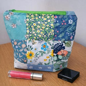 May include: A patchwork zipped pouch featuring various floral and botanical fabric squares in shades of blue, green, yellow, and white. The pouch has a bright green zip closure. A pink glitter lip gloss and a small black compact are nearby.
