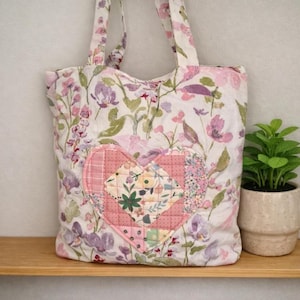 May include: A fabric tote bag featuring a floral pattern in shades of pink, purple, and green on a white background. The centre of the bag displays a quilted heart-shaped patch made from various patterned fabric squares in pink and floral prints.