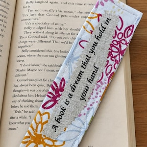 May include: A rectangular fabric bookmark featuring a white base with a colourful floral pattern in shades of pink, orange, and blue. A beige linen strip is stitched down the centre with the black text, A book is a dream that you hold in your hand.