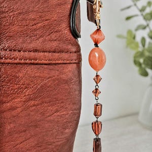 May include: A close-up of a brown leather bag with a decorative beaded charm. The charm features various orange and brown beads, including faceted and oval shapes, suspended from a gold-coloured clasp.