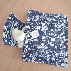 May include: A small, square fabric pouch featuring a dark navy blue background with a white floral and vine pattern. The pouch has a flap secured by a single white plastic snap button. Inside the pouch, a pair of white plastic earbuds are visible.