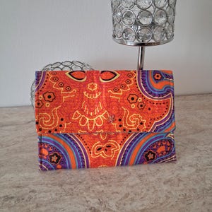 May include: An orange wallet with a paisley pattern in purple, blue, and yellow. The wallet is closed and has a flap closure. The background includes a decorative silver lamp.