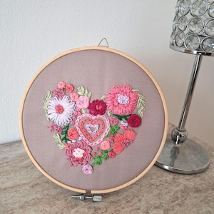 May include: A circular wooden embroidery hoop containing a taupe fabric panel. The fabric features a hand-stitched heart-shaped arrangement of various pink, red, and lavender flowers, leaves, and nested heart outlines in textured thread.