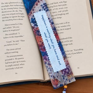 May include: A rectangular fabric bookmark with a blue, purple, and orange tie-dye pattern. A white centre strip features the text "Happiness is wanting what you have and not what you want" in blue. A beaded tassel hangs from the bottom edge.