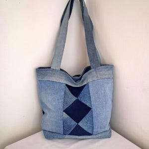 May include: A denim tote bag with a patchwork design. The bag features a diamond pattern in the centre, with lighter and darker shades of blue denim. The handles are made of the same denim material.