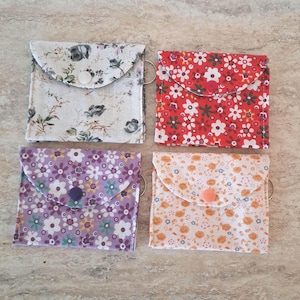 May include: Four square fabric pouches with snap closures and side rings. One is white with grey floral patterns, one is red with white and green flowers, one is purple with white and yellow flowers, and one is cream with small orange floral prints.