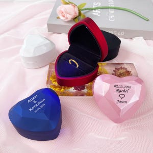 May include: A collection of heart-shaped ring boxes in various colors: white, blue, and pink. One box is open, revealing a gold ring. The pink box is engraved with a date and names. The blue box is engraved with names and a date.