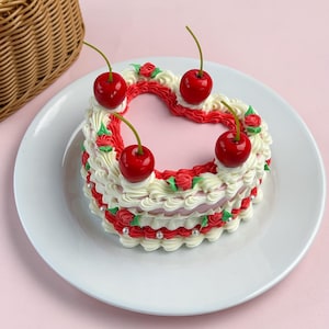 Op de afbeelding: Een hartvormige cake met witte frosting, rode accenten en vier kersen. De cake staat op een wit bord tegen een roze achtergrond. Een rieten mand is gedeeltelijk zichtbaar in de linkerbovenhoek.