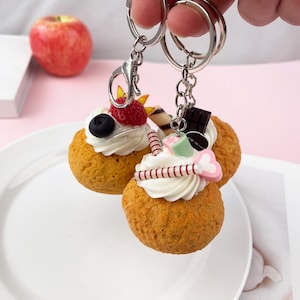 Pode incluir: Chaveiros com designs de sobremesas em miniatura. Cada chaveiro tem uma pequena base de bolo redonda coberta com glacê branco e elementos decorativos como frutas vermelhas, chocolate e um pingente em forma de coração rosa e branco. Os chaveiros são presos a argolas prateadas.