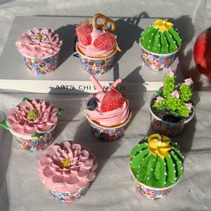 Puede incluir: Una colección de cupcakes decorativos con varios diseños. Algunos cupcakes están cubiertos con glaseado rosa y fresas, mientras que otros se asemejan a cactus con glaseado verde y flores amarillas. Los envoltorios de los cupcakes presentan un patrón colorido.