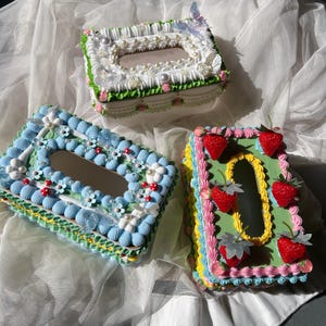 May include: Three decorative tissue box covers with ornate, colorful frosting-like designs. One is white with pearls and a butterfly, another is blue with flowers, and the third is green with strawberries.