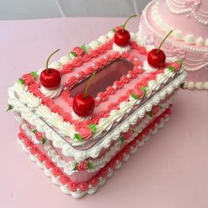 Puede incluir: Una caja de pañuelos rosa decorada con glaseado blanco y rosa, perlas falsas y cerezas rojas. La caja rectangular tiene una abertura rectangular y está diseñada para parecerse a un pastel. Un pastel rosa está en el fondo.