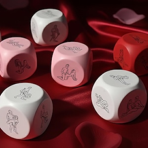 Può includere: Un gruppo di dadi in vari colori, tra cui bianco, rosa e rosso. Ogni dado presenta illustrazioni in nero di posizioni sessuali. I dadi sono disposti su una superficie di raso rosso.