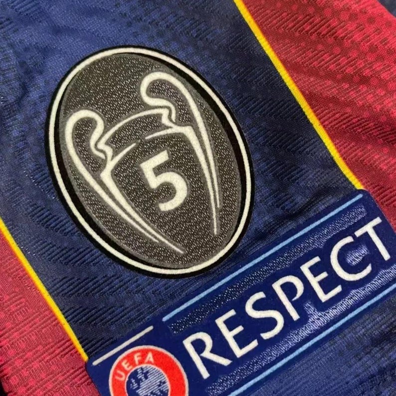 May include: Close-up of a blue and red sports jersey with a patch featuring the number 5 inside a trophy design. The jersey also has a "RESPECT" patch and a UEFA logo.
