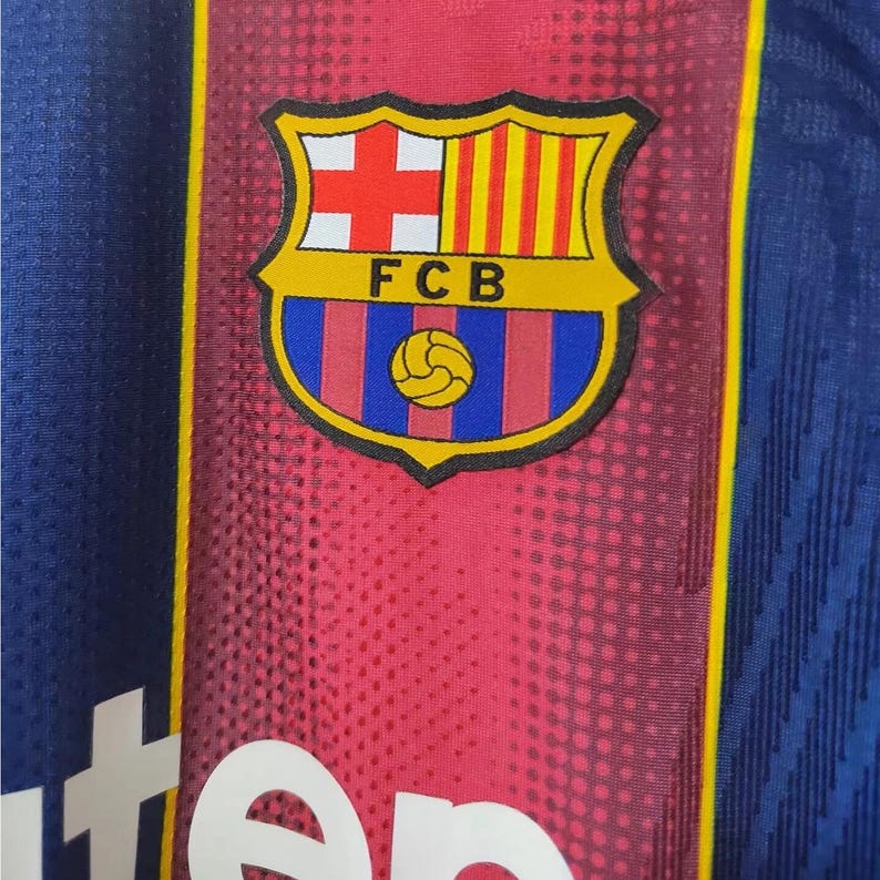 May include: Close-up of a blue and red jersey with the FCB emblem. The emblem features a shield with a cross, stripes, and a soccer ball. The jersey has a textured pattern and yellow accents.