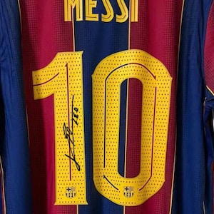 May include: A close-up of a Barcelona soccer jersey. The jersey is primarily maroon and blue with yellow accents. The number 10 is prominently displayed in yellow, with a signature below. The name "MESSI" is printed at the top.