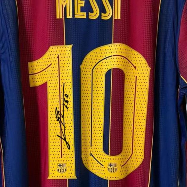 May include: A close-up of a Barcelona soccer jersey. The jersey is primarily maroon and blue with yellow accents. The number 10 is prominently displayed in yellow, with a signature below. The name "MESSI" is printed at the top.