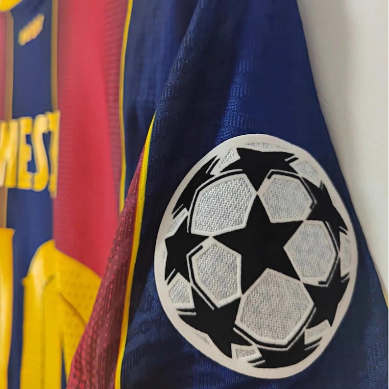 May include: Close-up of a blue, red, and yellow soccer jersey. The jersey features a white and black soccer ball patch on the sleeve. The jersey has a mesh texture and the word "WEST" is visible.