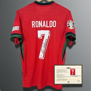Puede incluir: Camiseta de fútbol roja con el nombre "RONALDO" y el número "7" en la espalda. La camiseta tiene una firma debajo del número y un parche en la manga. Se muestra un certificado de autenticidad debajo.