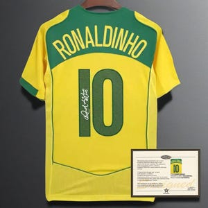 Puede incluir: Una camiseta de fútbol amarilla y verde con el nombre "RONALDINHO" y el número "10" en la espalda. La camiseta tiene una firma y un certificado de autenticidad.