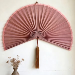 May include: A large, semi-circular wall fan in shades of pink, with a fringed edge. The fan is made of pleated material and has a brown tassel hanging from the center. The fan is mounted on a white wall.