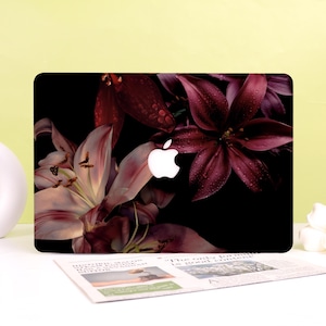 Op de afbeelding: Een laptopskin met een donker bloemendesign van diepbordeauxrode en lichtroze lelies tegen een zwarte achtergrond. De lelies zijn bedekt met kleine waterdruppels en in het midden van de skin bevindt zich een uitsparing voor het witte Apple-logo.