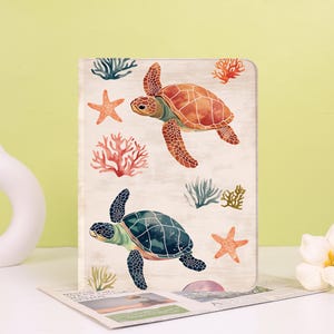 May include: A notebook with a watercolour design featuring two sea turtles, coral, and starfish. The turtles are in shades of orange and teal, with a light beige background. The notebook is open, revealing a glimpse of a newspaper.