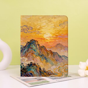 Puede incluir: Una funda rectangular para tableta con una pintura impresionista de una cadena montañosa al atardecer. La obra muestra vibrantes pinceladas amarillas, naranjas y azules que representan picos, nubes y un sol brillante en el cielo sobre las montañas.