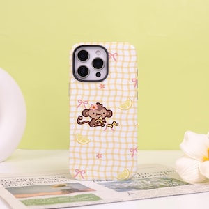May include: A smartphone case featuring a yellow and white checkered pattern with a cartoon monkey eating a banana, small pink bows, orange flowers, and yellow lemon slices scattered across the surface. The case has a black camera lens cutout.