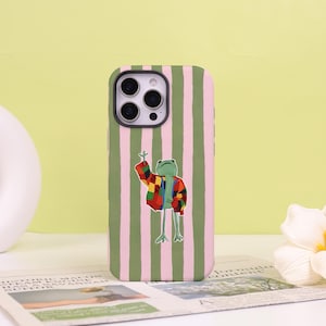 May include: A phone case featuring a vertical striped pattern in light green and pale pink. Centered on the case is an illustration of a green frog wearing a colorful patchwork jacket with red, yellow, blue, and black squares, waving one hand.