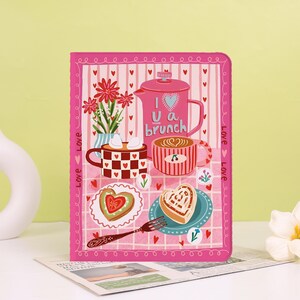 May include: A rectangular pink notebook featuring a graphic illustration of a French press, two mugs, heart-shaped pastries, and a fork. The text "I love brunch" is printed on the French press, and the word "love" repeats along the side borders.