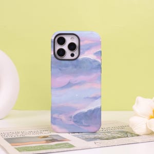 May include: A smartphone case with a pastel sky design. The case features a painted scene of fluffy clouds in shades of purple, pink, and blue. The camera cutouts are visible at the top. The case is on a white surface.